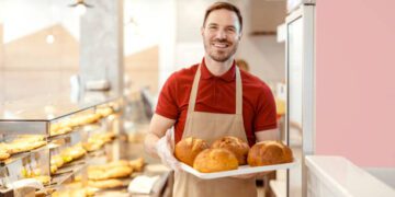Bakery shop owner with loaf of bred smiling to the camera while standing at pastry shop