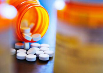 Pills pour out of a prescription medication bottle that rests on a table and is framed by two out of focus pill bottles.