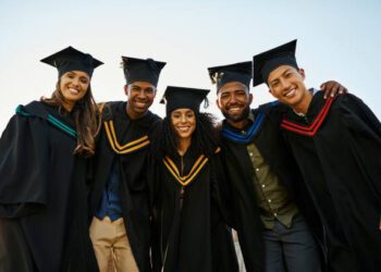 Portrait of diverse group of students in graduation gowns and caps with their arms around each other during graduation ceremony on university campus. Smiling friends standing close together at college